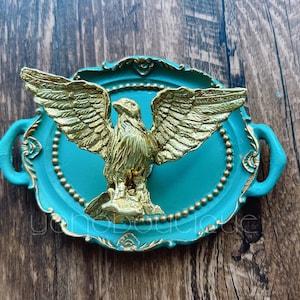 Pode incluir: Uma bandeja azul turquesa com detalhes dourados e uma figura de águia dourada no centro. A bandeja tem duas alças nas laterais.