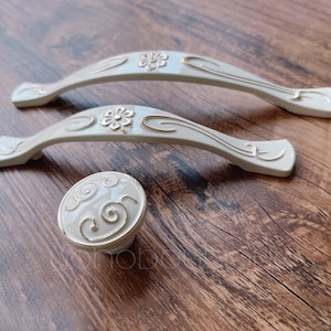 Peut inclure: Deux poignées de tiroir en céramique blanche avec des accents dorés et un seul bouton en céramique blanche avec des accents dorés.