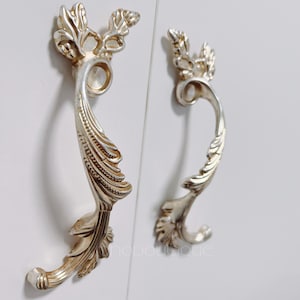May include: Two silver-toned decorative cabinet pulls with a floral design. The pulls have a curved shape and are accented with intricate details.