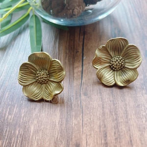 Könnte beinhalten: Zwei antike Schrankknöpfe in Blumenform aus Messing. Jeder Knopf hat detaillierte Blütenblatt- und Mittelteil-Designs. Die Knöpfe werden auf einer Holzoberfläche präsentiert, was auf ihre Verwendung in der Möbelrestaurierung hindeutet.