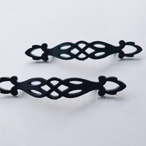 Könnte beinhalten: Zwei schwarze Metall-Schubladengriffe mit einem komplizierten, gewebten Design. Die Griffe haben einen dekorativen, verzierten Stil.