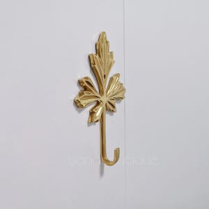 Op de afbeelding: Een gouden haak in de vorm van een blad met een decoratief ontwerp. De haak is gemonteerd op een witte muur.