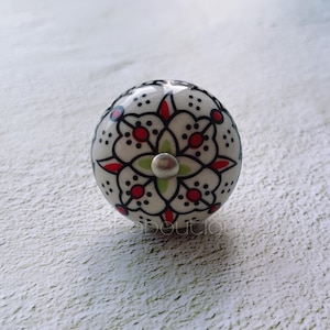 Op de afbeelding: Witte keramische knop met een zwart en rood bloemenmotief. De knop heeft een zilveren metalen schroef in het midden.