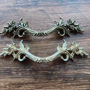 May include: Two ornate gold-toned metal drawer pulls with a decorative floral design.