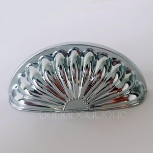 Może przedstawiać: Błyszczący, chromowany uchwyt do szafki o wachlarzowatym kształcie. Okucia są półkoliste i mają szereg zakrzywionych grzbietów promieniujących z centralnego punktu. Tekst "yonoboutique" jest widoczny na dole.