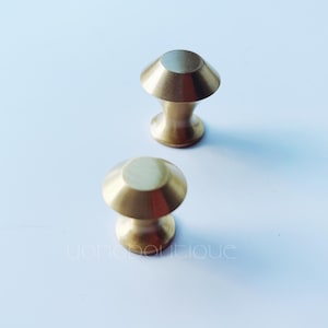 Könnte beinhalten: Zwei goldfarbene Schrankknöpfe mit abgerundeter, leicht gewölbter Oberseite und einem kurzen, zylindrischen Sockel.