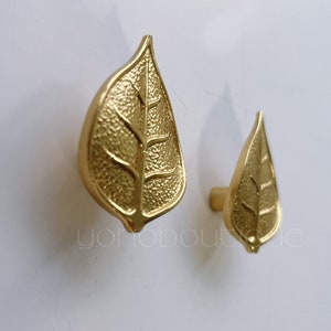 Może przedstawiać: Dwa złote gałki do szafek w kształcie liści. Większa gałka znajduje się po lewej stronie, a mniejsza po prawej.
