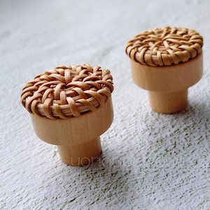 May include: Two wooden cabinet knobs with woven rattan tops. The knobs have a light brown finish and a cylindrical base. The woven tops are circular and feature a natural, light brown color. The knobs are set against a white background.