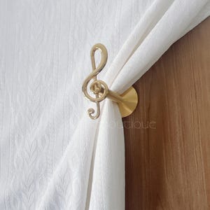 May include: A brass-coloured curtain tieback shaped like a treble clef. It is holding a white curtain. This decorative item adds a musical theme to window treatments and is a stylish home decor accessory.