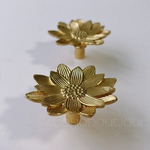 May include: Two gold-toned metal cabinet knobs shaped like stylized sunflowers. The knobs have a raised, textured design and a smooth, round base.
