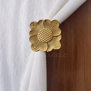 Puede incluir: Un recogedor de cortinas dorado en forma de flor. El recogedor está hecho de metal y tiene un diseño floral detallado. Los pétalos están en capas y el centro tiene un patrón texturizado. El recogedor sujeta una cortina blanca.