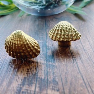 以下が含まれることがあります： 木製の表面に置かれた、2つの金色の貝殻型のキャビネットノブ。1つは完全な貝殻で、もう1つはキノコ型のノブです。ノブはテクスチャ加工された表面で、おそらく金属製です。