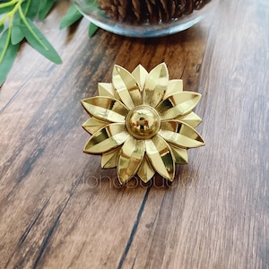 Könnte beinhalten: Ein goldfarbener, metallener Knopf in Blütenform mit einem runden Zentrum. Der Knopf befindet sich auf einer Holzoberfläche.
