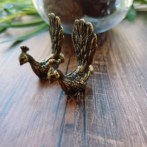 Puede incluir: Dos figuras de pavo real de bronce con diseños detallados de plumas. Los pavos reales están posados sobre una superficie de madera, con un globo de cristal en el fondo. Las figuras están intrincadamente detalladas y tienen una estética vintage.