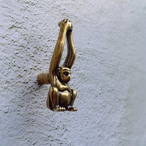 May include: A brass monkey-shaped cabinet knob. The knob features a sitting monkey with its arms raised. The monkey is a warm brass color and is mounted on a textured wall. The knob is designed for furniture.