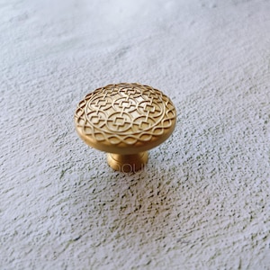 May include: A gold-toned cabinet knob with a circular, intricate design. The knob has a raised, geometric pattern.