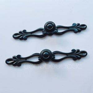 May include: Two black metal drawer pulls with ornate designs and round knobs. The pulls have a vintage style.
