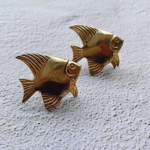 May include: Two gold-colored metal fish ornaments with a detailed design. The fish are facing opposite directions and have a slightly curved shape.