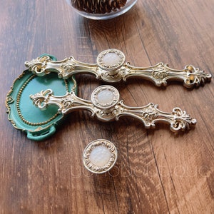 May include: Two ornate gold drawer pulls with white porcelain knobs. The pulls have a decorative scroll design and are shown on a wooden surface.