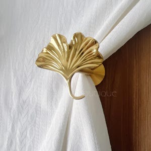 May include: A gold-colored, leaf-shaped curtain tieback. The tieback is made of metal and has a hook to hold the curtain in place. The curtain is white and has a textured pattern. The background is a wooden door.