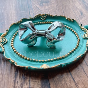 Könnte beinhalten: Ein dekoratives Tablett in Türkisblau mit goldenen Akzenten und einem Perlenrand. Das Tablett hat zwei Griffe und in der Mitte befindet sich eine silberne Schleife.