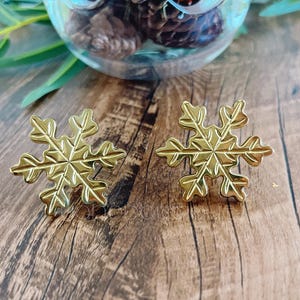 May include: Two gold-coloured snowflake-shaped stud earrings are displayed on a wooden surface. The earrings have intricate cutouts, and are positioned near a glass container filled with pinecones. Green leaves are visible in the background.