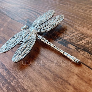 以下が含まれることがあります： 羽に複雑な模様が施された銀色のトンボ。トンボは木製の表面にとまっている。