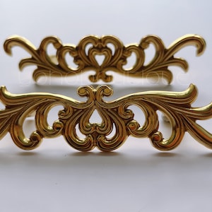 Puede incluir: Dos tiradores de cajón de metal dorado con un diseño decorativo de desplazamiento.