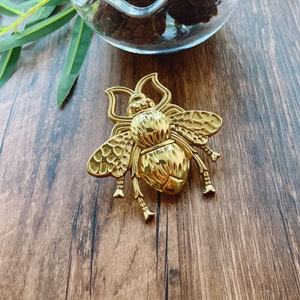 以下が含まれることがあります： 複雑なディテールが施された金色の蜂のブローチ。蜂は質感のある体と羽を持ち、脚は丸まっています。