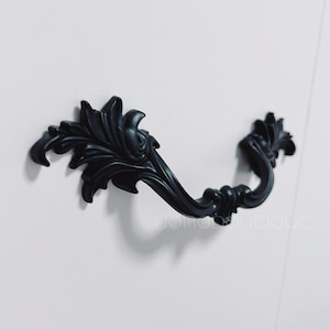 Peut inclure: Une poignée de meuble noire et ornée, avec un design incurvé. La poignée présente un motif floral détaillé et est montée sur une surface blanche. Ce matériel est un élément décoratif pour les meubles.
