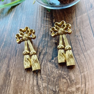 Puede incluir: Un par de pendientes de metal dorado con un diseño de lazo y detalles de borla.