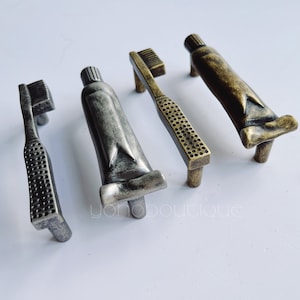 May include: Four antique-style cabinet knobs in the shape of toothbrushes and toothpaste tubes. The knobs are made of metal and have a distressed finish.