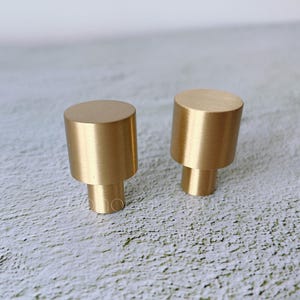 Pode incluir: Dois botões de armário de metal dourado com forma cilíndrica. Os botões são lisos e têm um acabamento polido.
