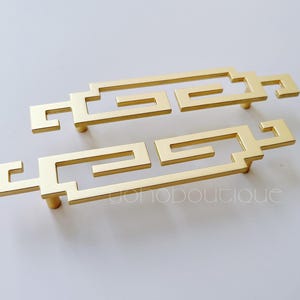 May include: Two gold-toned cabinet pulls with a geometric design. The pulls are made of metal and have a smooth finish.