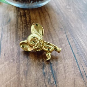 Op de afbeelding: Een gouden metalen pin in de vorm van een cartoonmuis. De muis staat in een speelse pose met zijn oren omhoog en zijn staart gekruld.