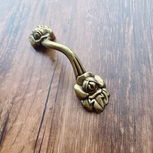 Könnte beinhalten: Ein goldfarbener Metall-Schubladengriff mit Rosenmuster an beiden Enden. Der Griff ist gebogen und hat eine glatte Oberfläche.