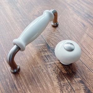 Könnte beinhalten: Weißer Keramik-Schrankgriff und passender Knopf mit gebürsteter Nickel-Hardware. Der Griff ist im Stangenstil mit gebogenen Enden. Der Knopf ist rund mit einer flachen, gebürsteten Nickel-Oberseite. Auf einer Holzmaserung.