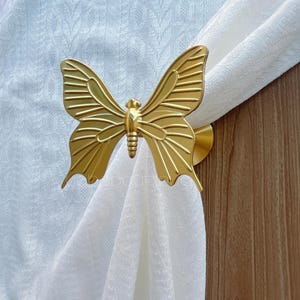 May include: A gold-coloured butterfly curtain tieback. The butterfly design features detailed wing patterns and is holding a white curtain. The tieback is mounted on a wooden surface.