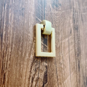 May include: Gold square cabinet pull with a rounded top. The pull is mounted on a wood surface.