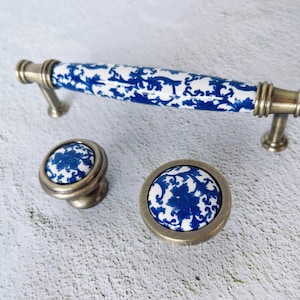 Peut inclure: Un ensemble de quincaillerie d'armoire comprenant une longue poignée et deux boutons. La quincaillerie présente une finition en laiton et des inserts en céramique blanche avec des motifs floraux bleus.