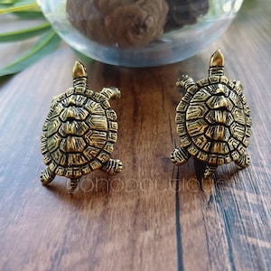 Op de afbeelding: Twee antieke goudkleurige schildpadfiguren. Elke schildpad heeft een gedetailleerd schildpatroon, vier kleine pootjes en een kop met een kleine vin. De figuren worden op een houten oppervlak gepresenteerd.