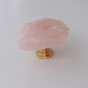 Op de afbeelding: Een rozenkwarts kristallen knop met een messing voet. Het roze kristal heeft een ruwe, natuurlijke textuur. De knop is een decoratief accent voor meubels of kasten. De tekst "yohoboutique" is op het oppervlak gedrukt.