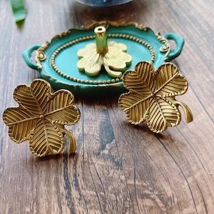 May include: Two gold-colored four-leaf clover shaped decorative pieces on a wooden surface. The clovers have a textured design and are sitting on a green tray.
