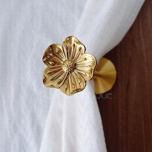 Könnte beinhalten: Ein goldfarbener Metall-Gardinenhalter in Form einer Blume. Der Halter hält einen weißen Vorhang. Die Blume hat detaillierte Blütenblätter und ein strukturiertes Zentrum. Das Wort "yonboutique" ist unten im Bild zu sehen.