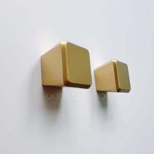 Peut inclure: Deux boutons de cabinet carrés dorés, l'un plus grand que l'autre, sur un fond blanc.