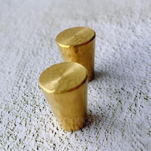 Op de afbeelding: Twee gouden gehamerde metalen kastknoppen. De knoppen zijn rond en hebben een getextureerd oppervlak.