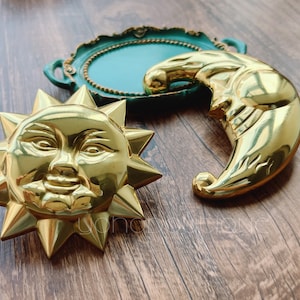 Pode incluir: Peças decorativas douradas de sol e lua com traços faciais detalhados. O sol tem raios pontiagudos e a lua tem formato de meia-lua. Uma bandeja decorativa turquesa com acabamento dourado está ao fundo.