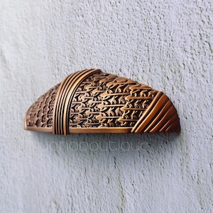 Puede incluir: Un tirador de armario de tono cobre con un intrincado diseño tejido. El tirador tiene forma de semicírculo y un acabado liso y pulido.