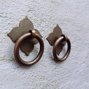 May include: Two brass drawer pulls with a floral design and a ring handle. The pulls are mounted on a white wall.