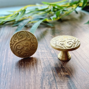 Pode incluir: Dois botões de armário dourados com um design ornamentado e em espiral. O botão à esquerda é plano, enquanto o botão à direita tem uma base elevada.